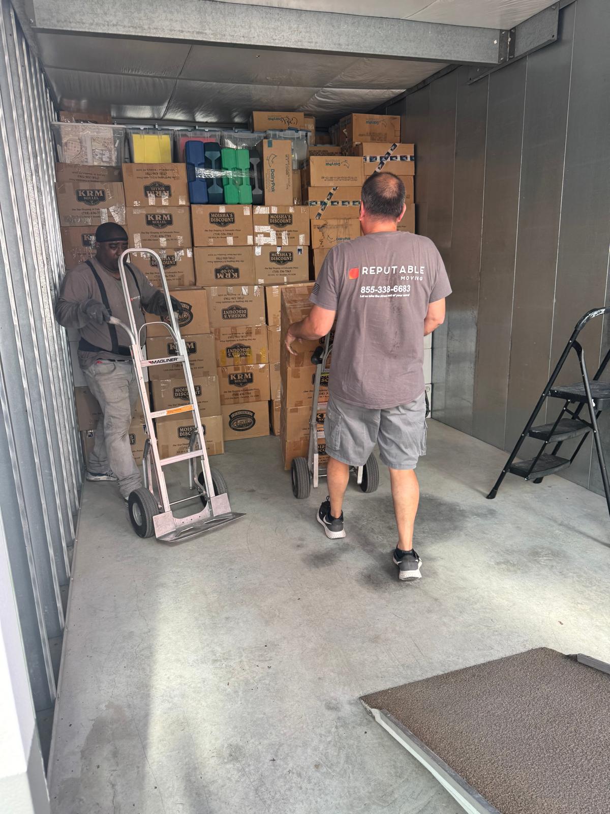 Two crew with hand trucks inside a Reputable storage unit packed wall-to-wall with branded boxes