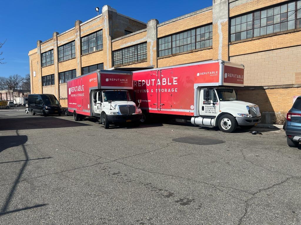 Two red Reputable Moving and Storage trucks parked tight against an industrial brick warehouse wall